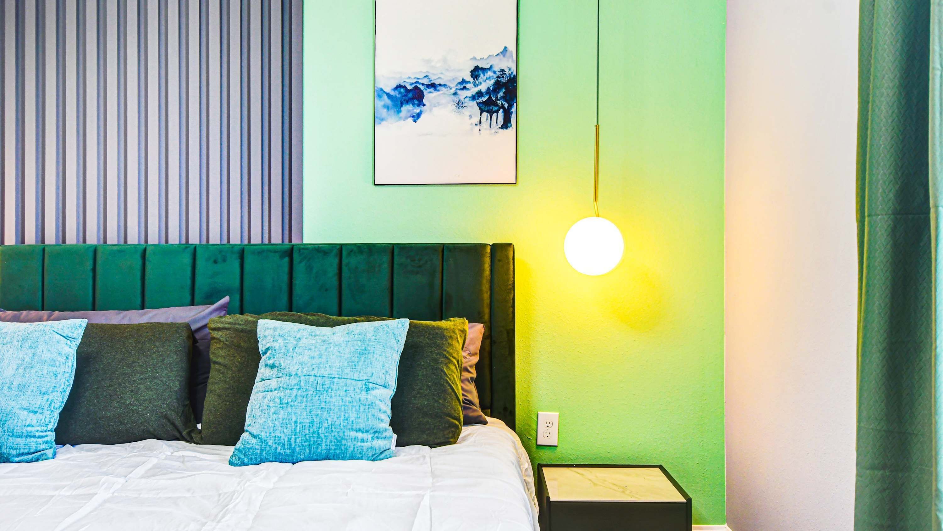 A bed with a green headboard, multiple pillows, a bedside table, and a wall-mounted light.