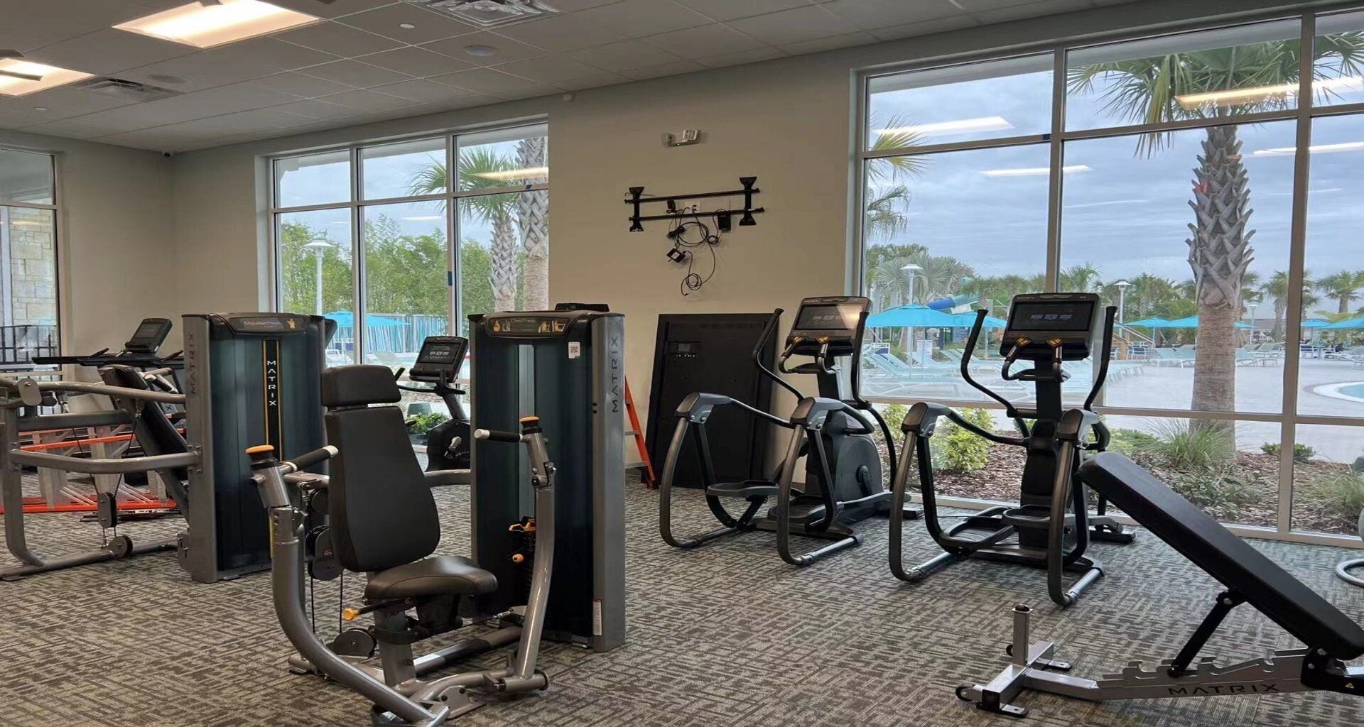 Een fitnessruimte met diverse trainingsapparaten, grote ramen en uitzicht op palmbomen en een zwembad.