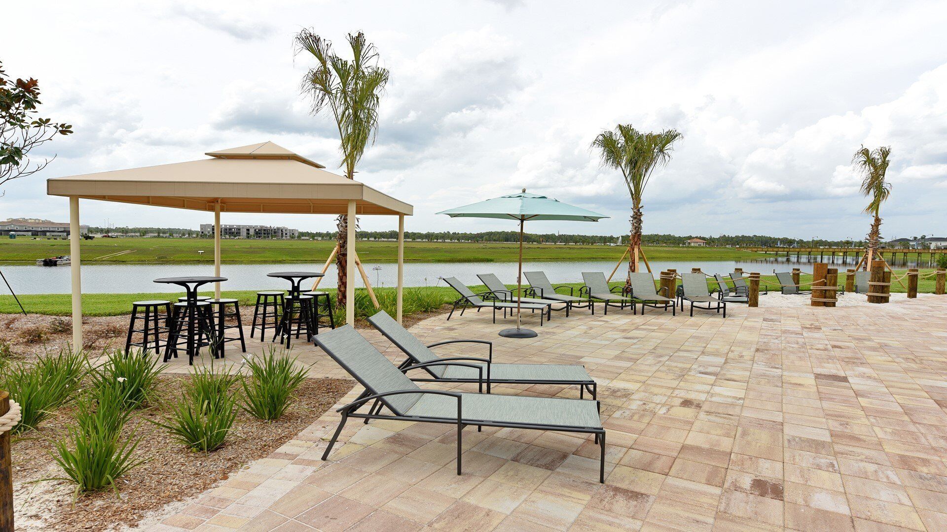 An outdoor patio with lounge chairs, tables, and umbrellas, overlooking a body of water and a distant building.