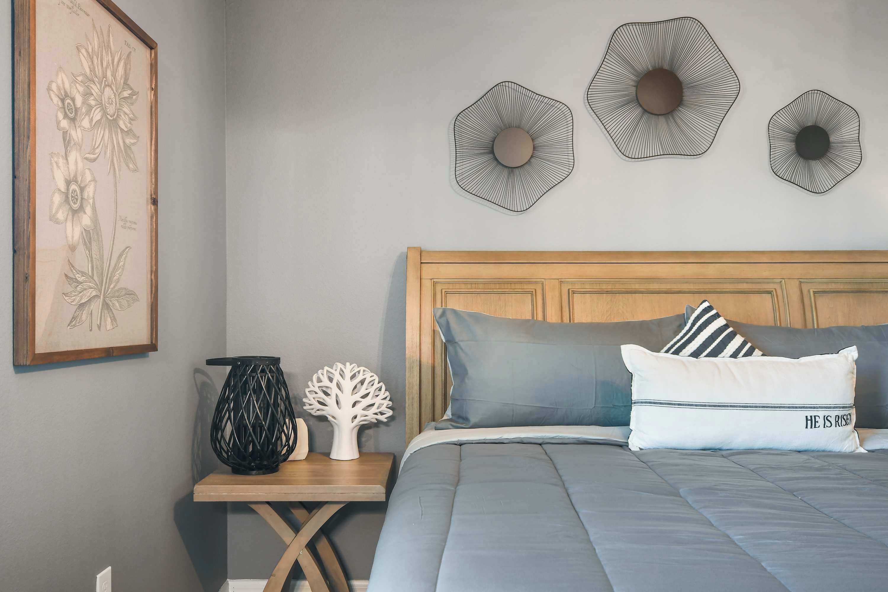 A bedroom with a wooden headboard, a bed with grey bedding, a framed botanical illustration, and three wall-mounted decorative items.