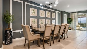 A dining room with a large table and six chairs, framed botanical prints on the wall, and a potted plant.