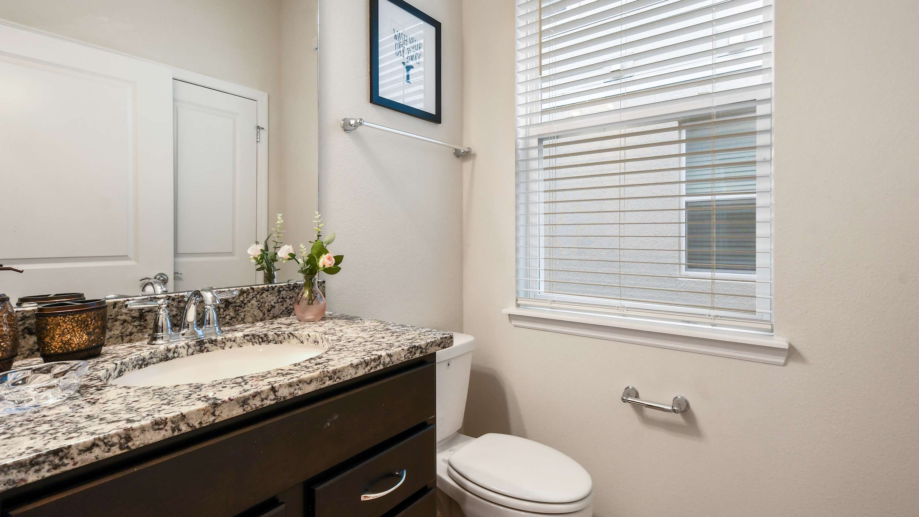 A bathroom with a granite countertop sink, a white toilet, and a window with blinds.