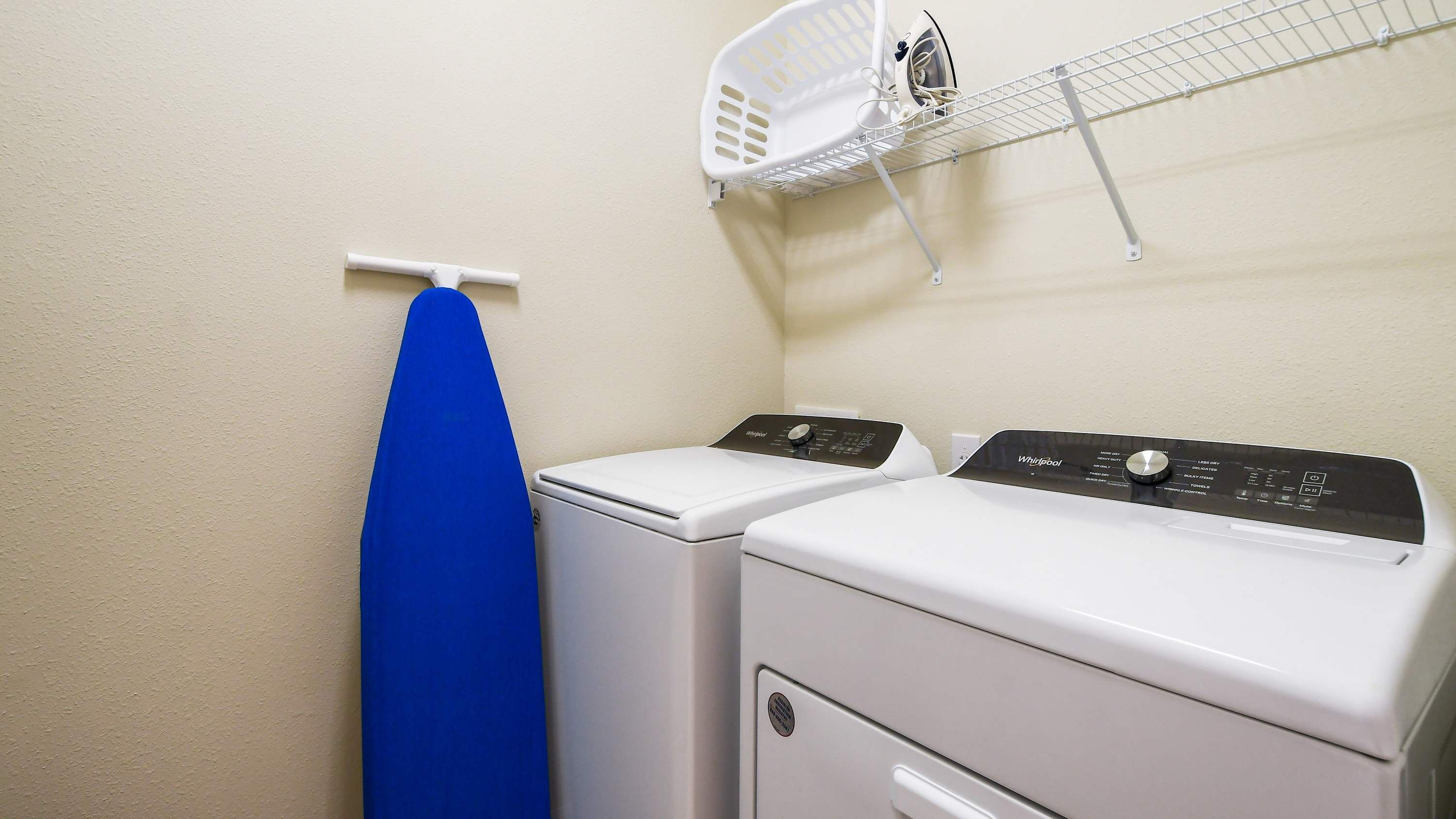 A laundry room with two white washers and dryers, a blue ironing board, and a white laundry basket on a wire shelf.
