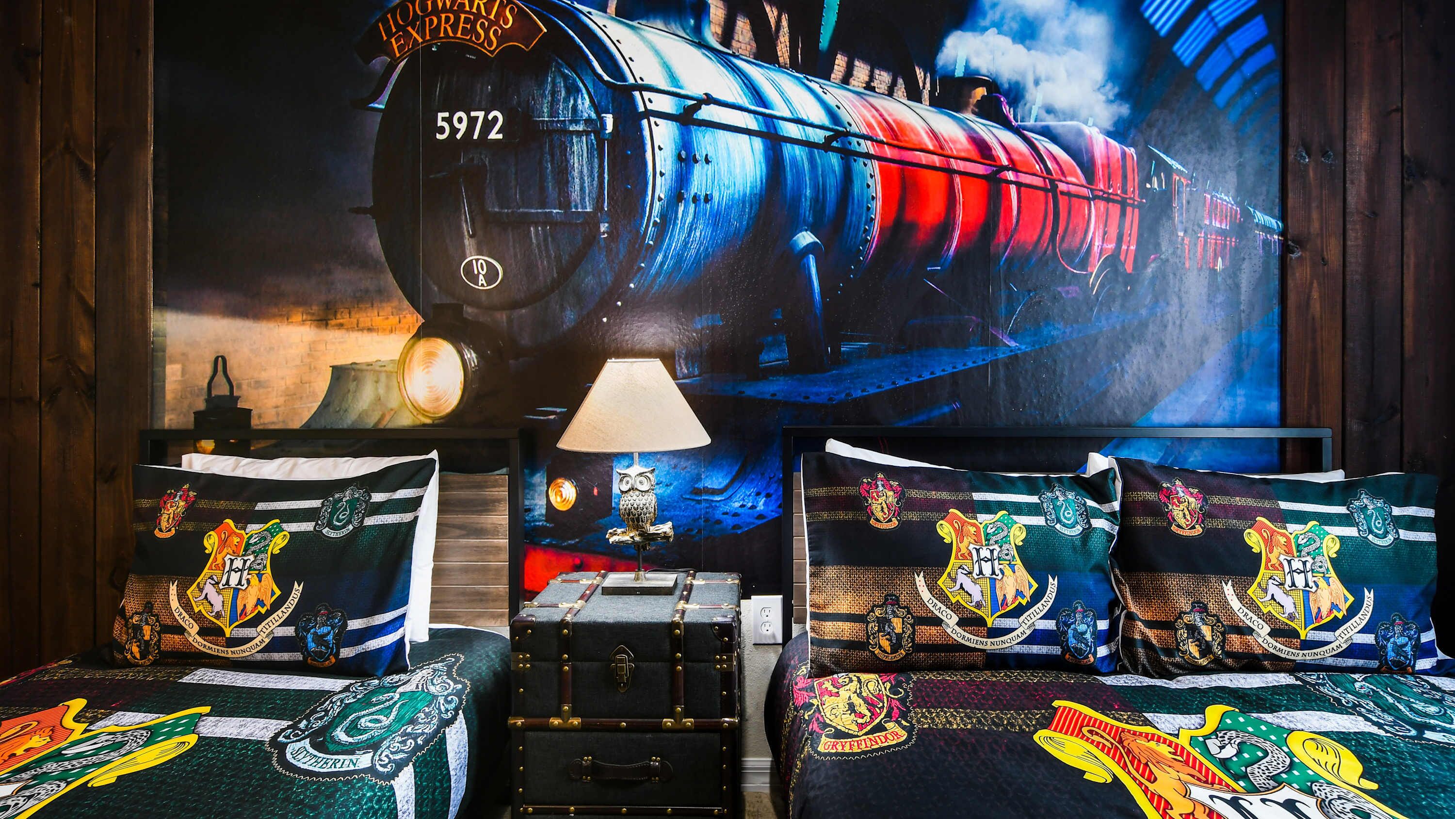 A room with a large Hogwarts Express poster, a lamp, a suitcase, and pillows with Hogwarts emblems.