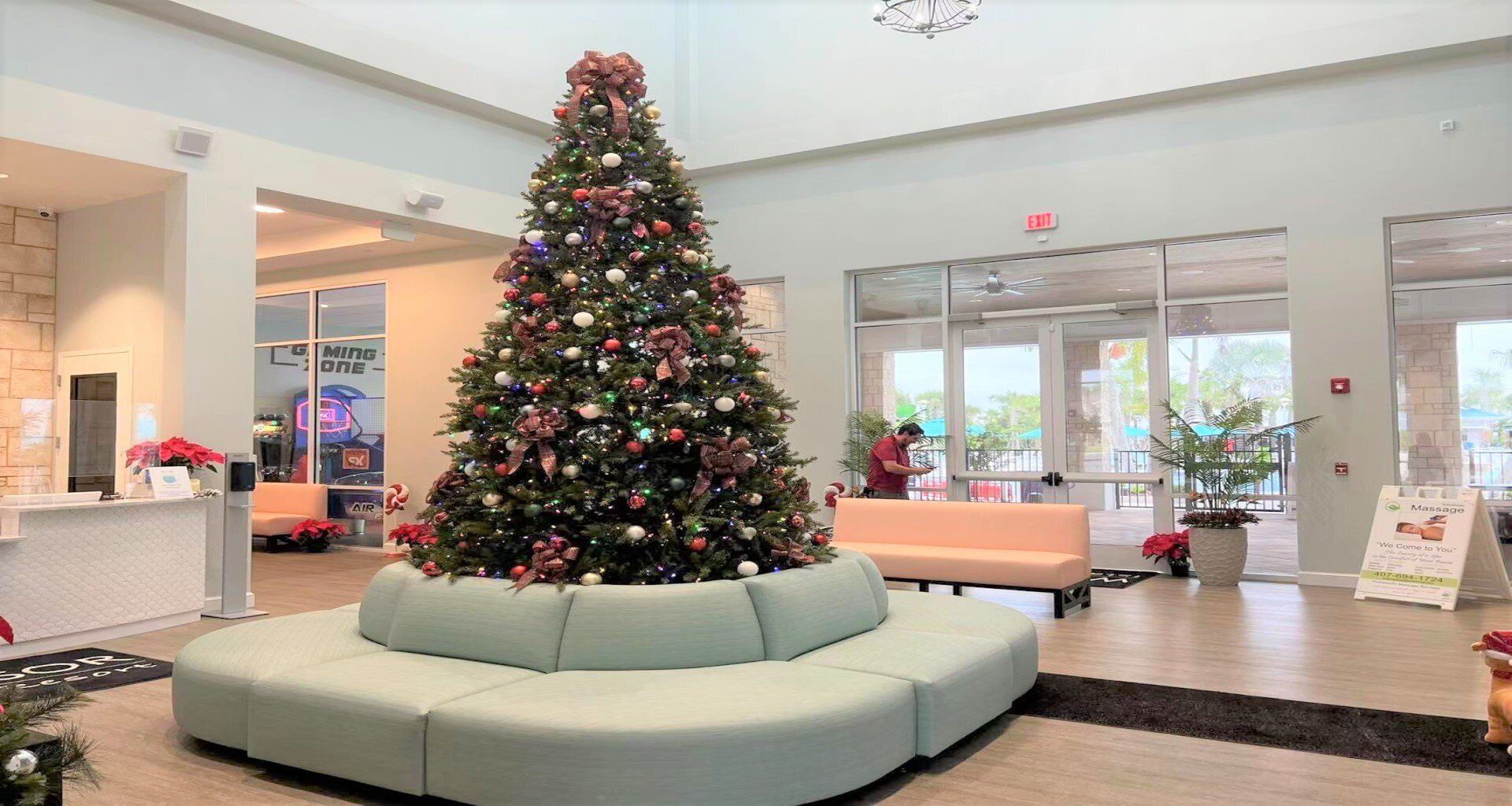 Un sapin de Noël dans un hall d’entrée, avec un canapé de couleur claire et un panneau indiquant un service de massage.