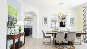 Un coin repas avec une table en bois, des chaises à revêtement gris et un lustre. On y trouve un vase vert avec une lampe sur une console, ainsi qu’un tableau encadré au mur.