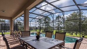 Un patio couvert, équipé d’une table et de chaises, donnant sur une piscine et un jardin aménagé.