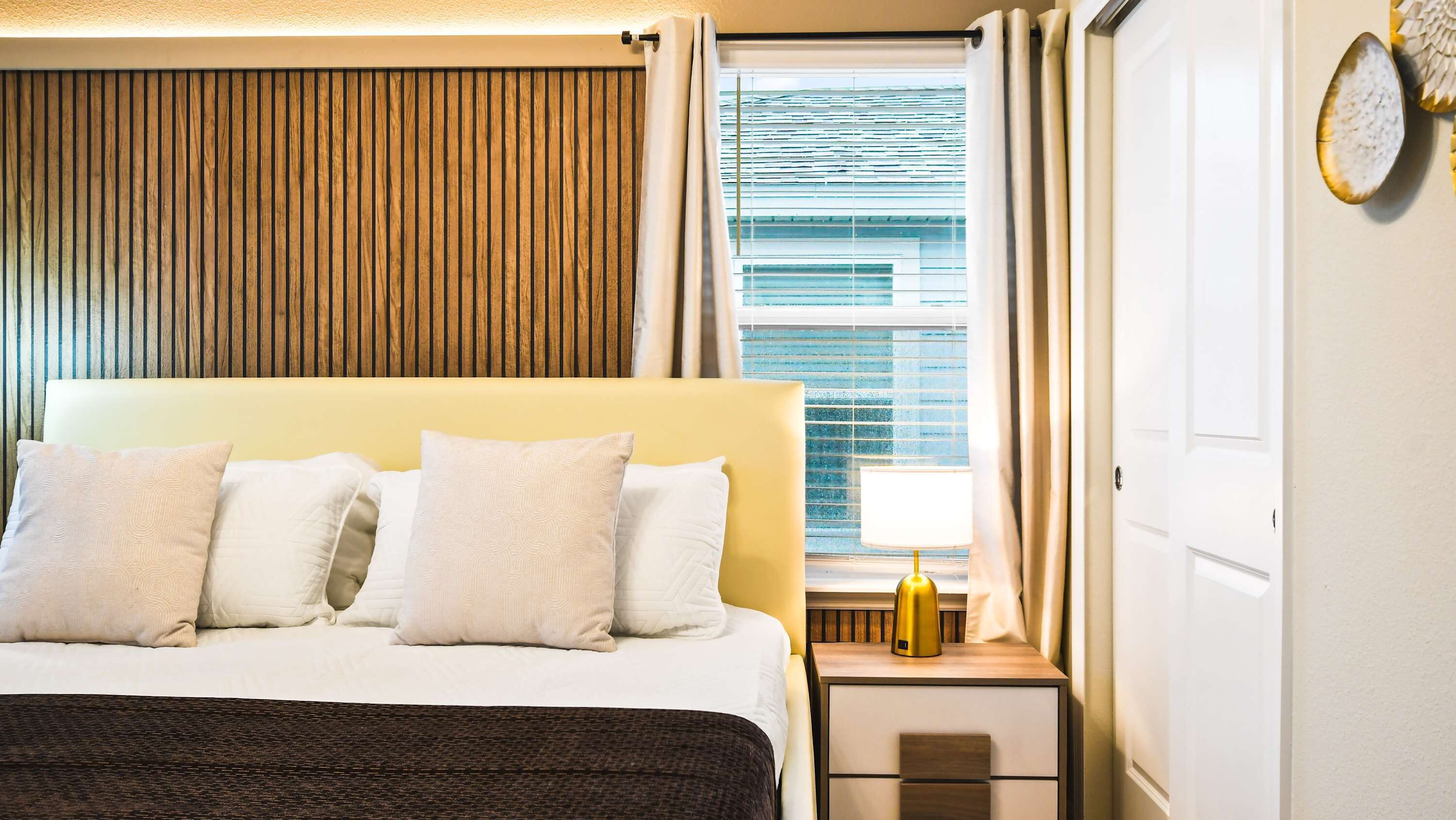 Quarto de hotel com cama, mesa de cabeceira, janela com persianas e porta.
