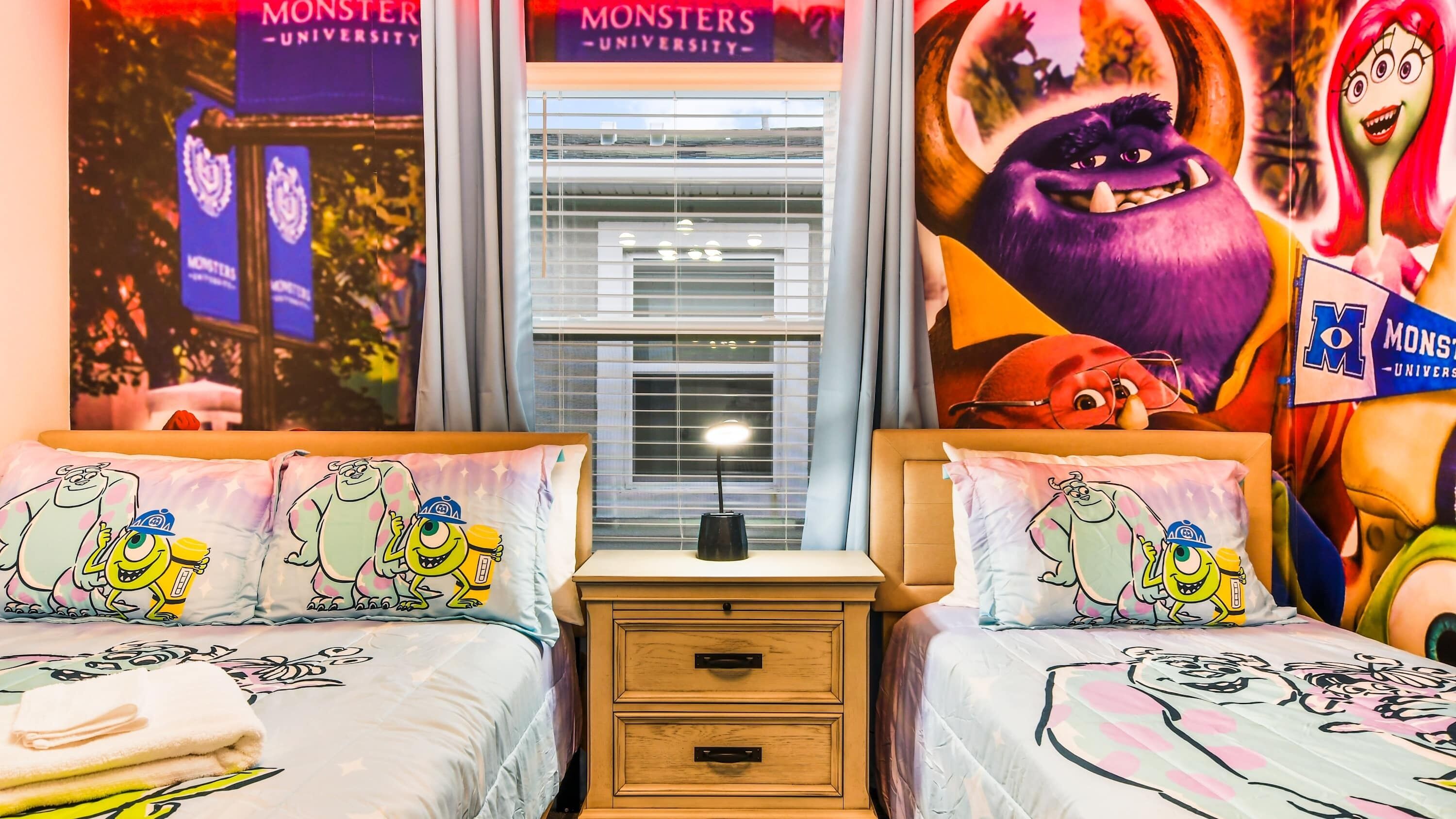 A bedroom with two beds, each with a Monsters University themed duvet, a bedside table with a lamp, and a window with blinds.