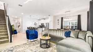 Un salon moderne avec un canapé gris, un fauteuil bleu et une petite table ronde. Un escalier se trouve à gauche et une cuisine est visible au fond.