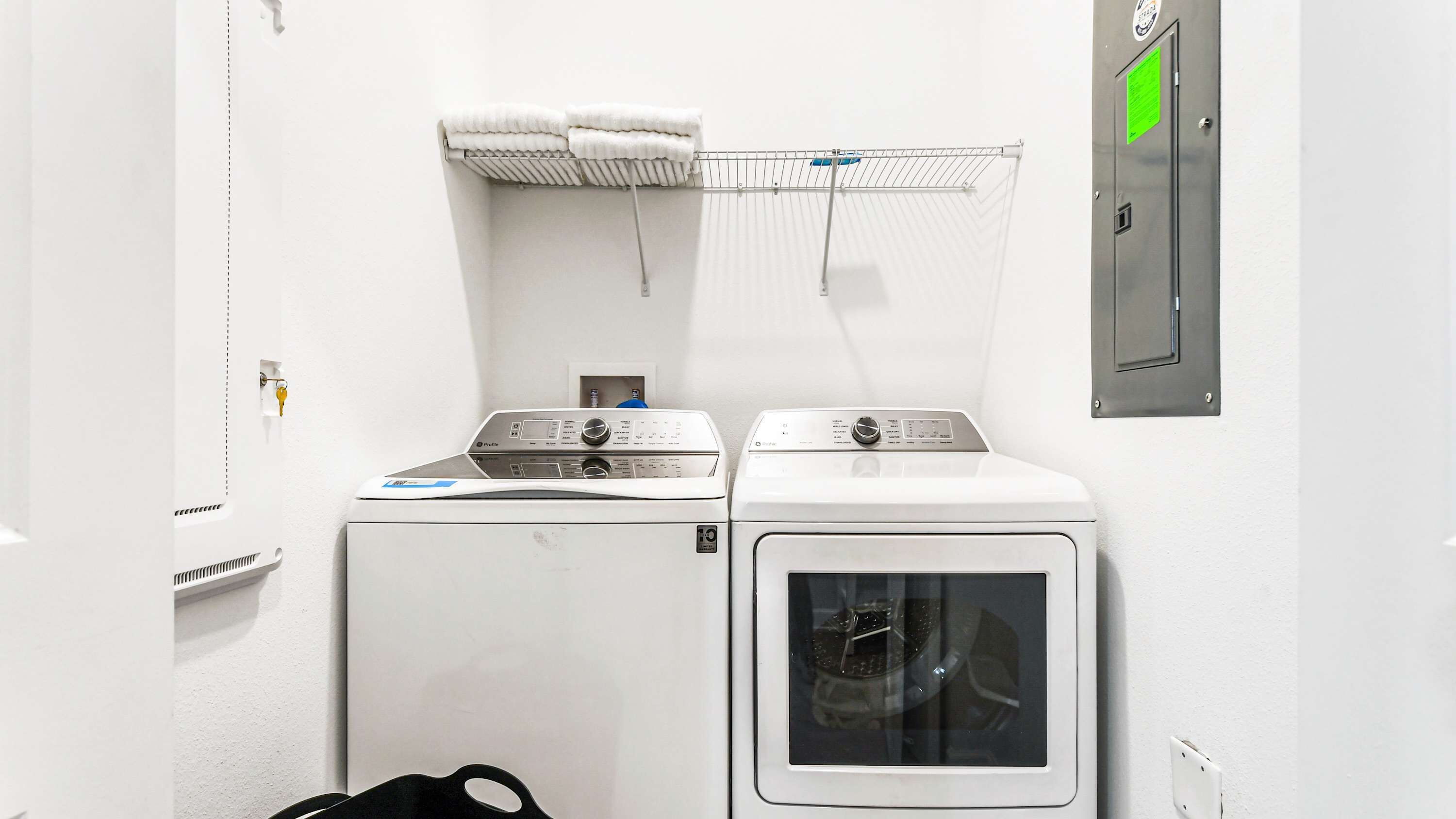 A laundry room with two washing machines and a dryer, a metal rack with folded towels, and a green sign on the wall.