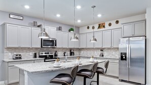 Uma cozinha moderna com armários brancos, eletrodomésticos de aço inoxidável, uma ilha de mármore com banquetas, iluminação pendente e uma placa de boas-vindas.