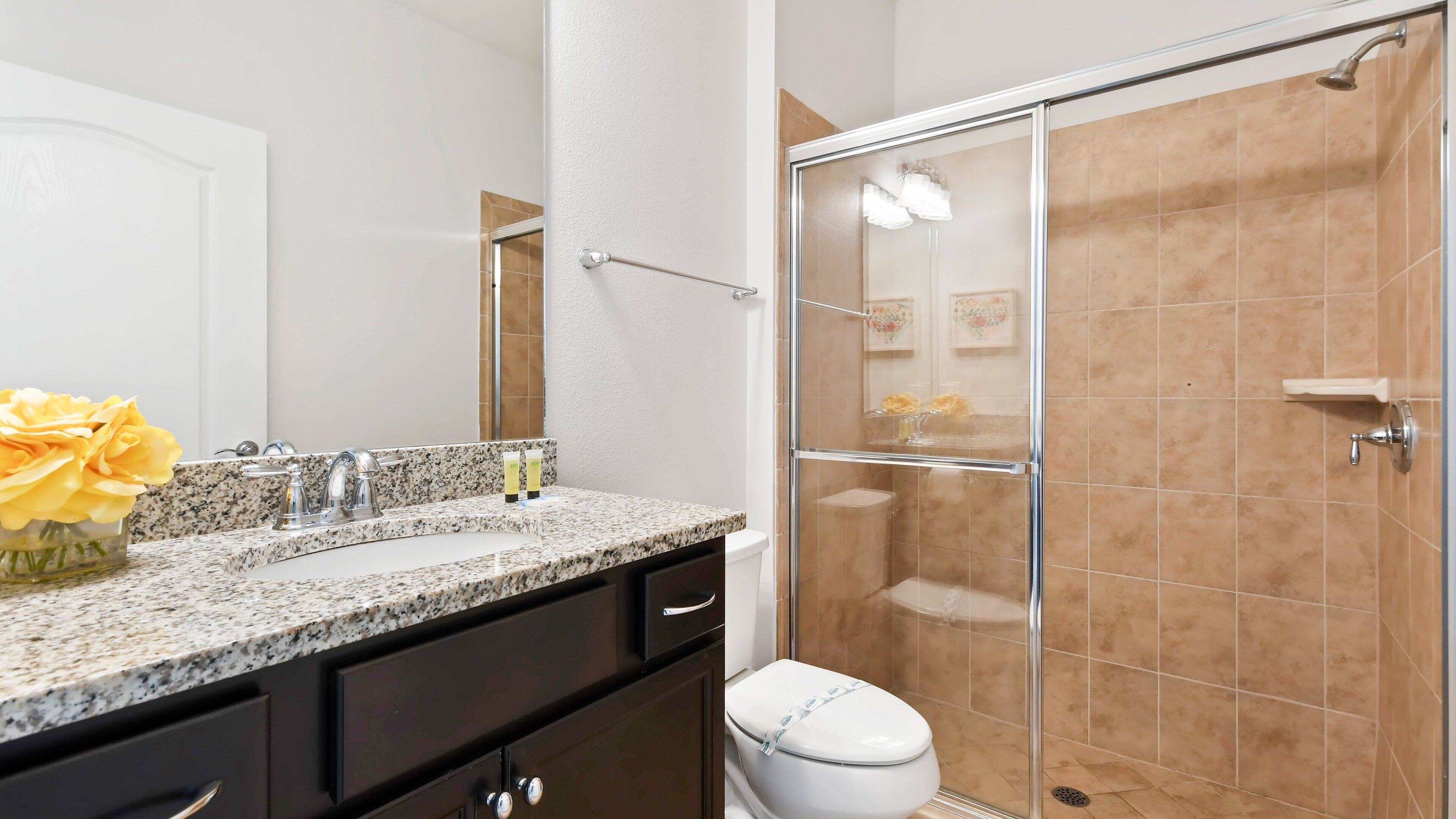 Un baño con encimera de granito, inodoro blanco, ducha con mampara de vidrio y espejo sobre el lavabo.