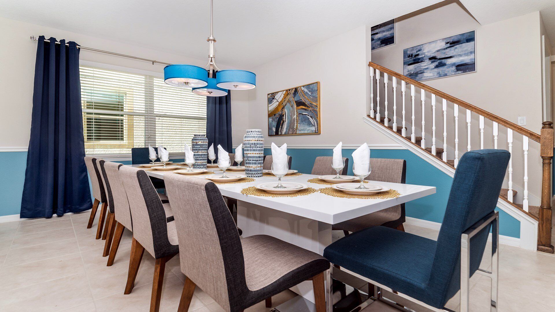 Een eetgedeelte met een witte tafel gedekt voor zes personen, blauwe stoelen en een trap met witte leuningen.