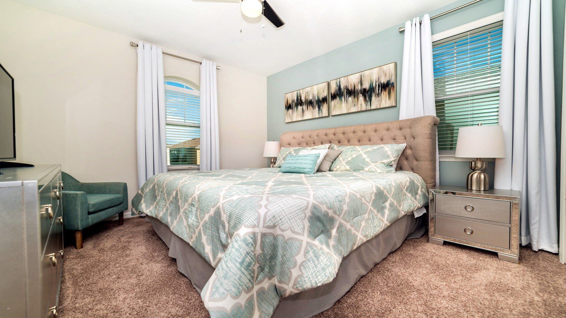Une chambre à coucher avec une tête de lit capitonnée, un lit avec une literie à motifs, une table de chevet avec une lampe, une chaise et une commode.