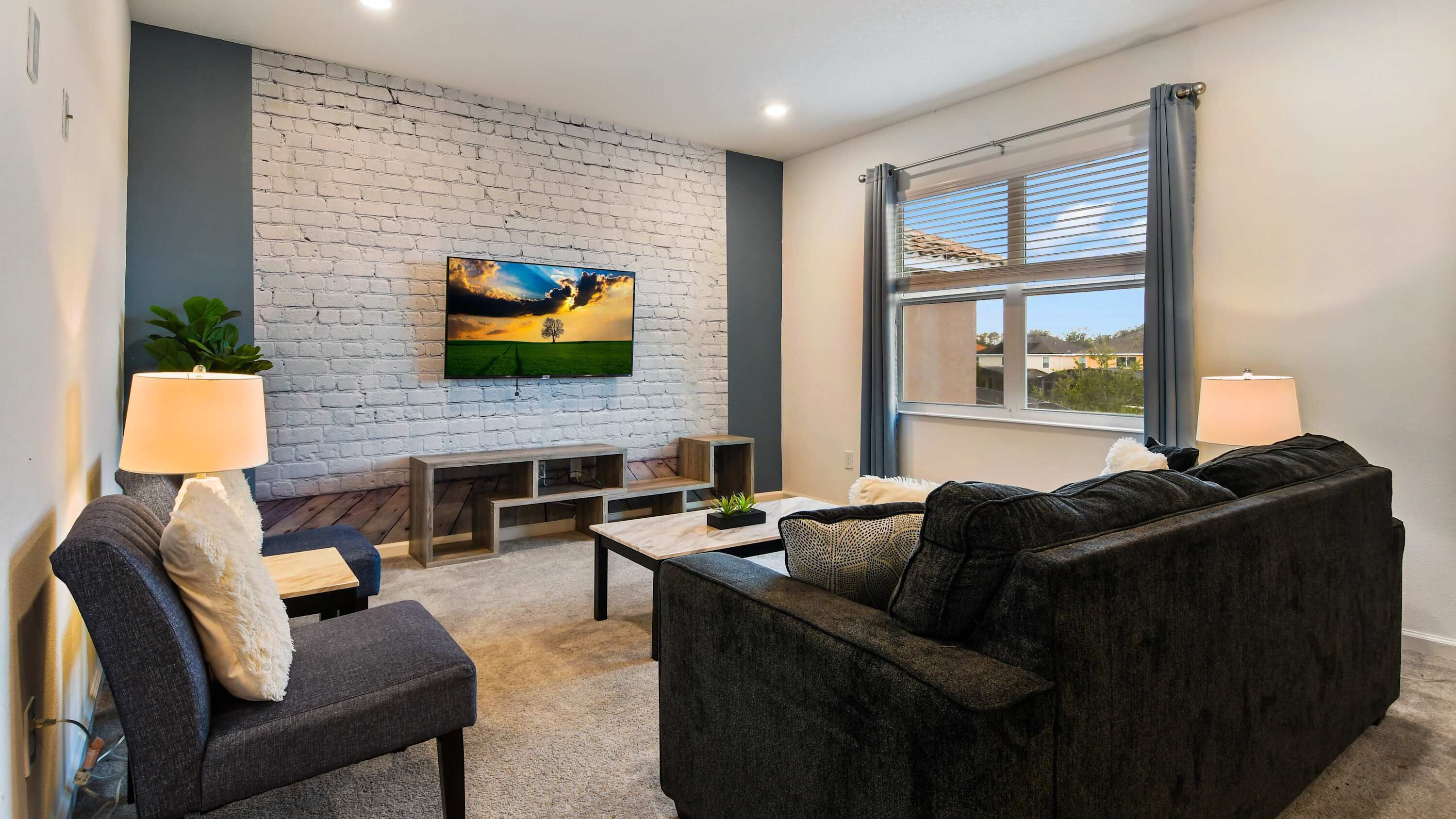 Uma sala de estar moderna com uma parede de tijolos brancos, uma televisão de tela plana grande, um sofá cinza escuro, duas poltronas, uma mesa de centro e uma janela com persianas.