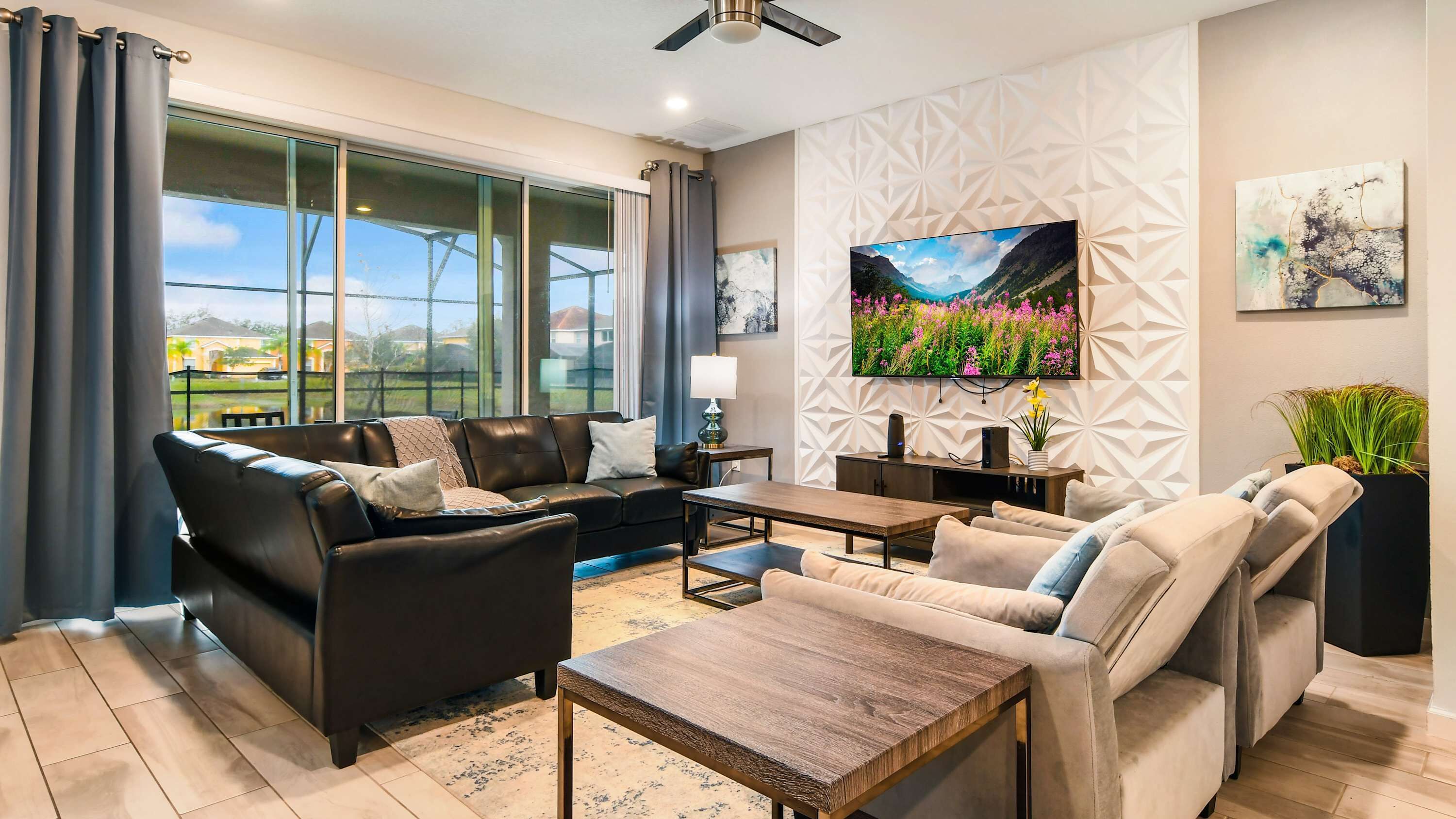 A modern living room with a sofa set, a coffee table, a TV mounted on the wall, and a large window offering a view of the outdoors.