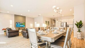 A modern dining area with a large table set for six, surrounded by cushioned chairs. A television is mounted on the wall, and there is a plant in the corner. - Spacious 12-bed Villa With Pool & Resort Amenities (Kissimmee)