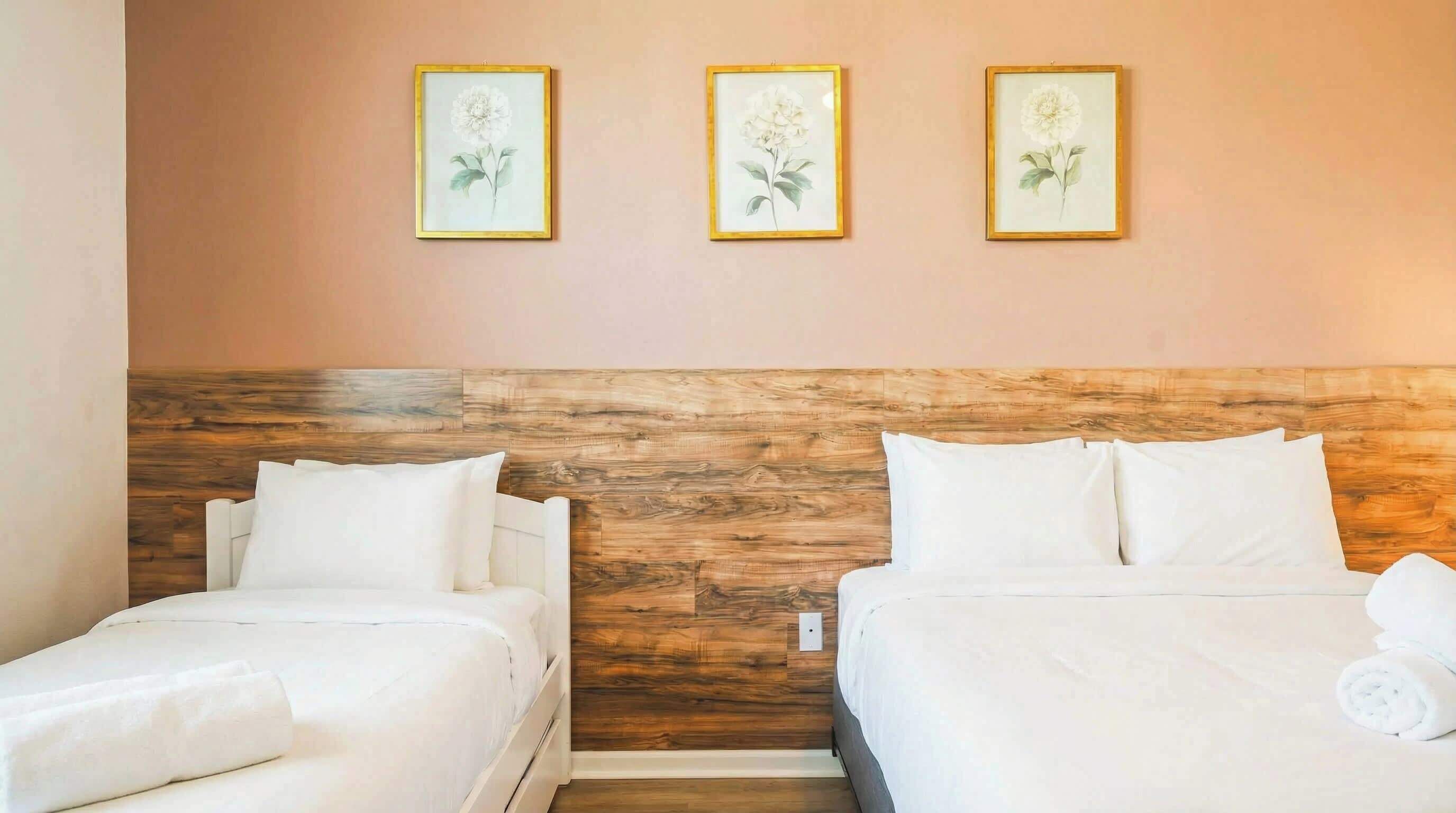 Quarto de hotel com duas camas, uma cabeceira de madeira e três quadros florais na parede.