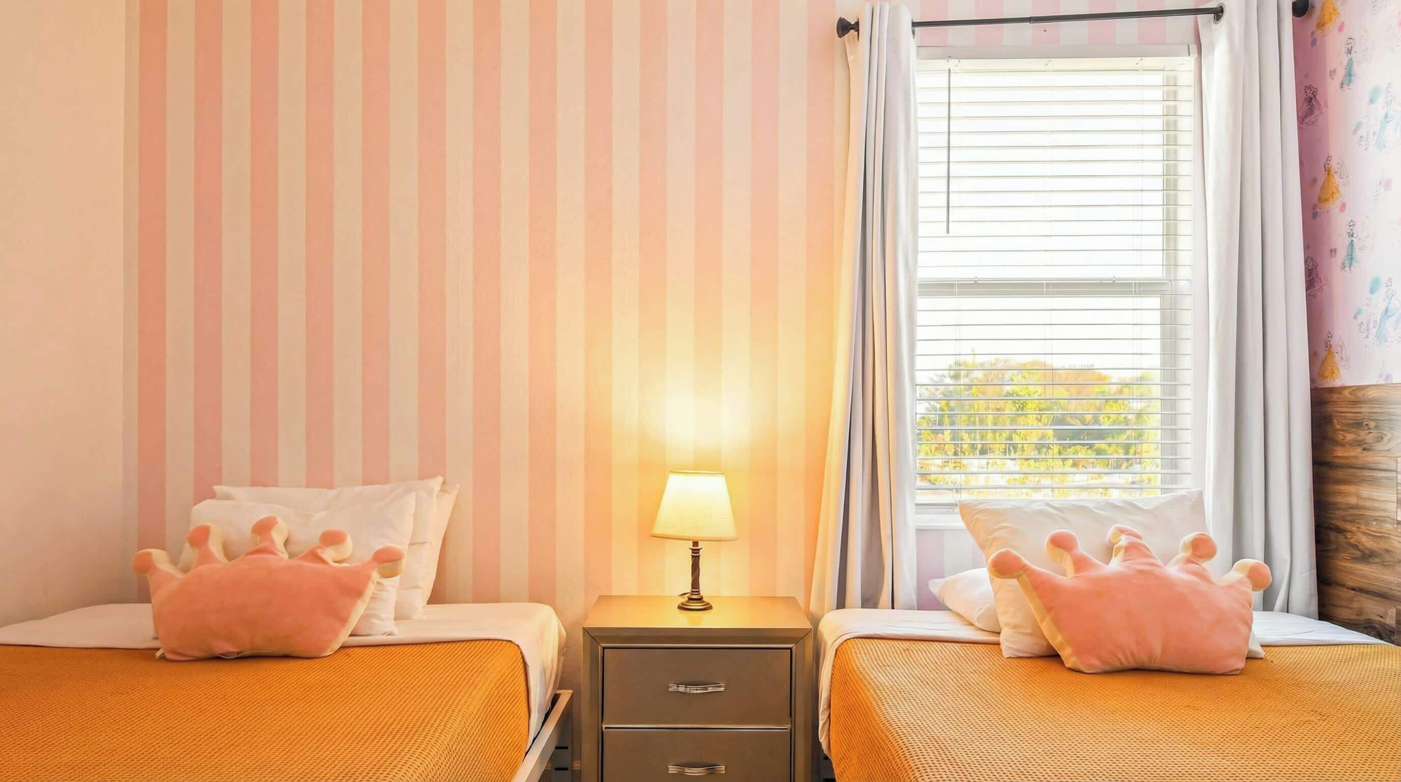 A room with two beds, each with a pink pillow, a bedside table with a lamp, and a window with blinds.