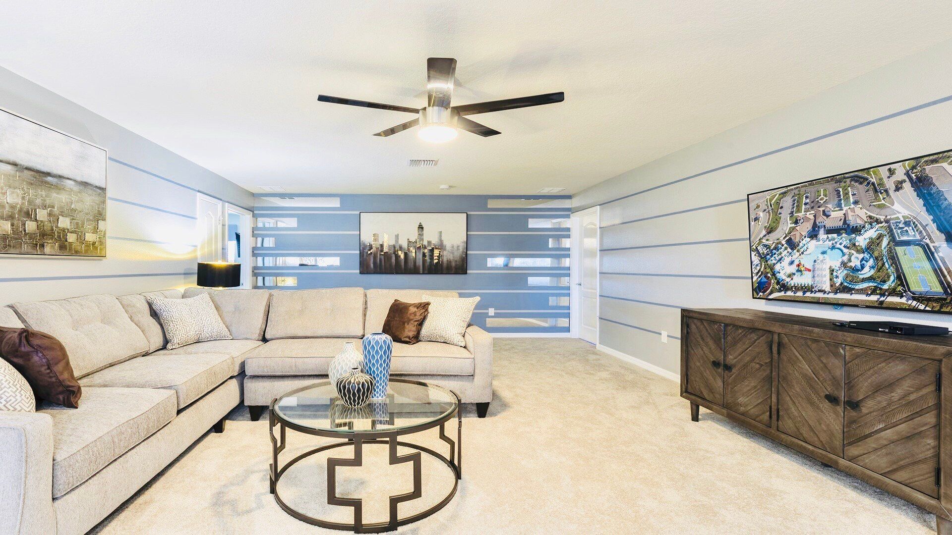 A living room with a sectional sofa, a glass coffee table, a ceiling fan, and a large TV on a wooden cabinet.