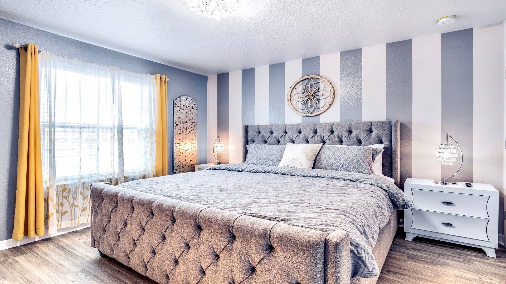 A bedroom with a tufted headboard, striped walls, a large window with curtains, a bedside table with a lamp, and a decorative wall piece.