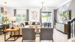 A modern living room with a sofa, two armchairs, a coffee table, and a dining area with a table and chairs. There is a TV mounted on the wall, a staircase with a railing, and a large window with curtains. - Storey Lake 5br Family Townhome w Pool Near Disney (Kissimmee)