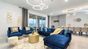 A modern living room with a blue sofa set, a gold coffee table, and a dining area with a white table and chairs. - Luxury Family Escape at Windsor Island Orlando (Davenport)