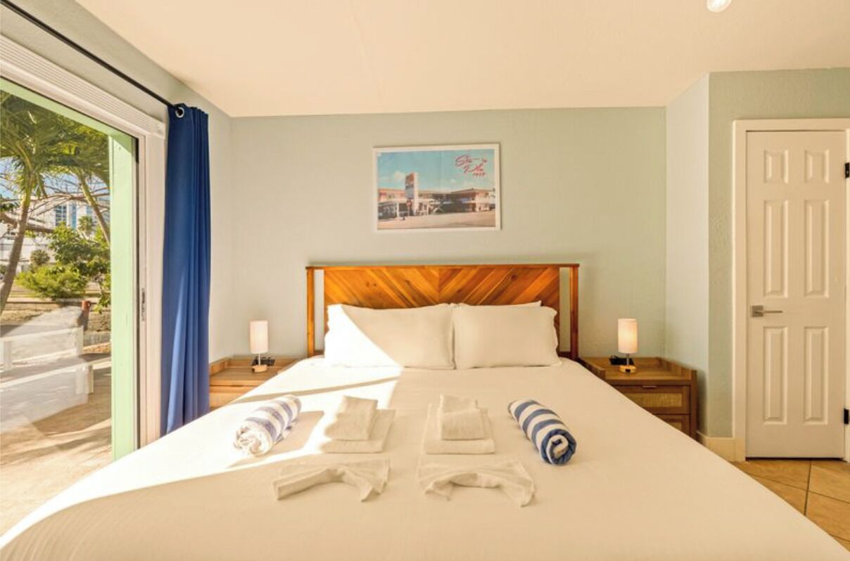 A hotel room with a large bed, two bedside tables with lamps, a painting on the wall, and a view of palm trees through a sliding door.
