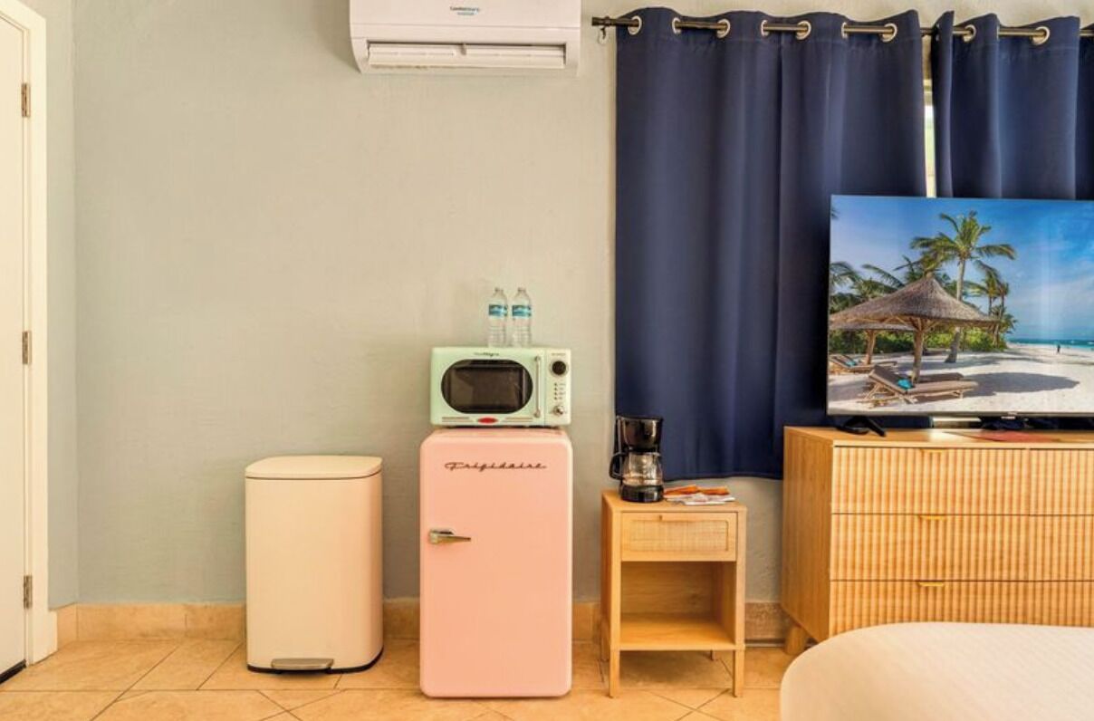 Une chambre d’hôtel compacte comprenant un lit, un téléviseur diffusant des images de plage, un minibar, une petite table avec une machine à café, une poubelle et un réfrigérateur.