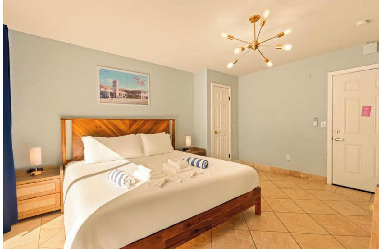 Quarto de hotel com cama de madeira, mesa de cabeceira com abajur, luminária no teto e porta com uma placa.