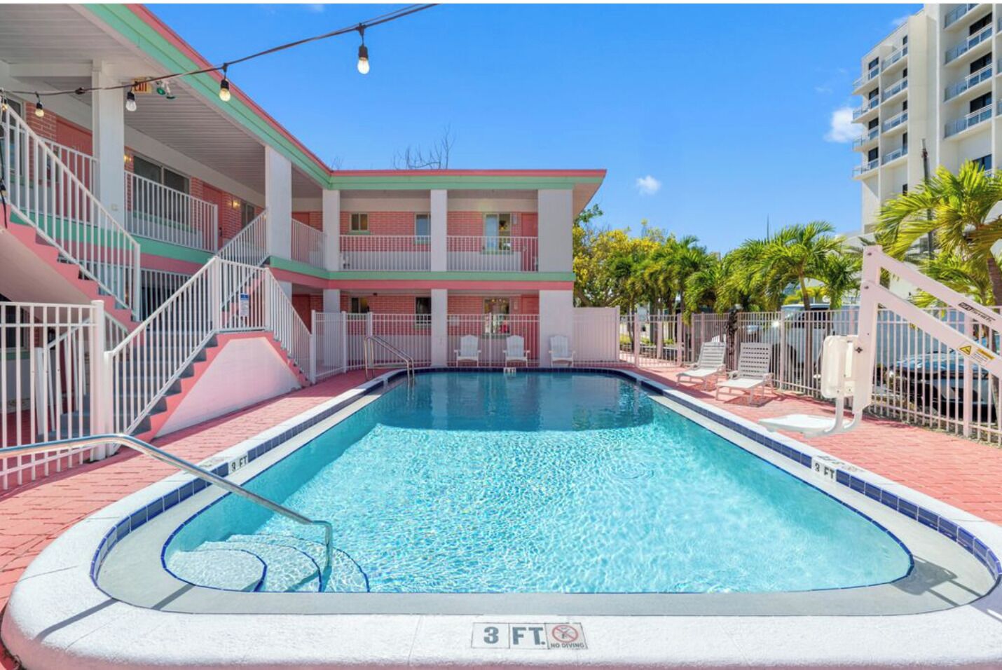 Una piscina con una marca de profundidad de 3 pies, rodeada por una valla blanca, con un edificio de fachada roja y blanca al fondo.