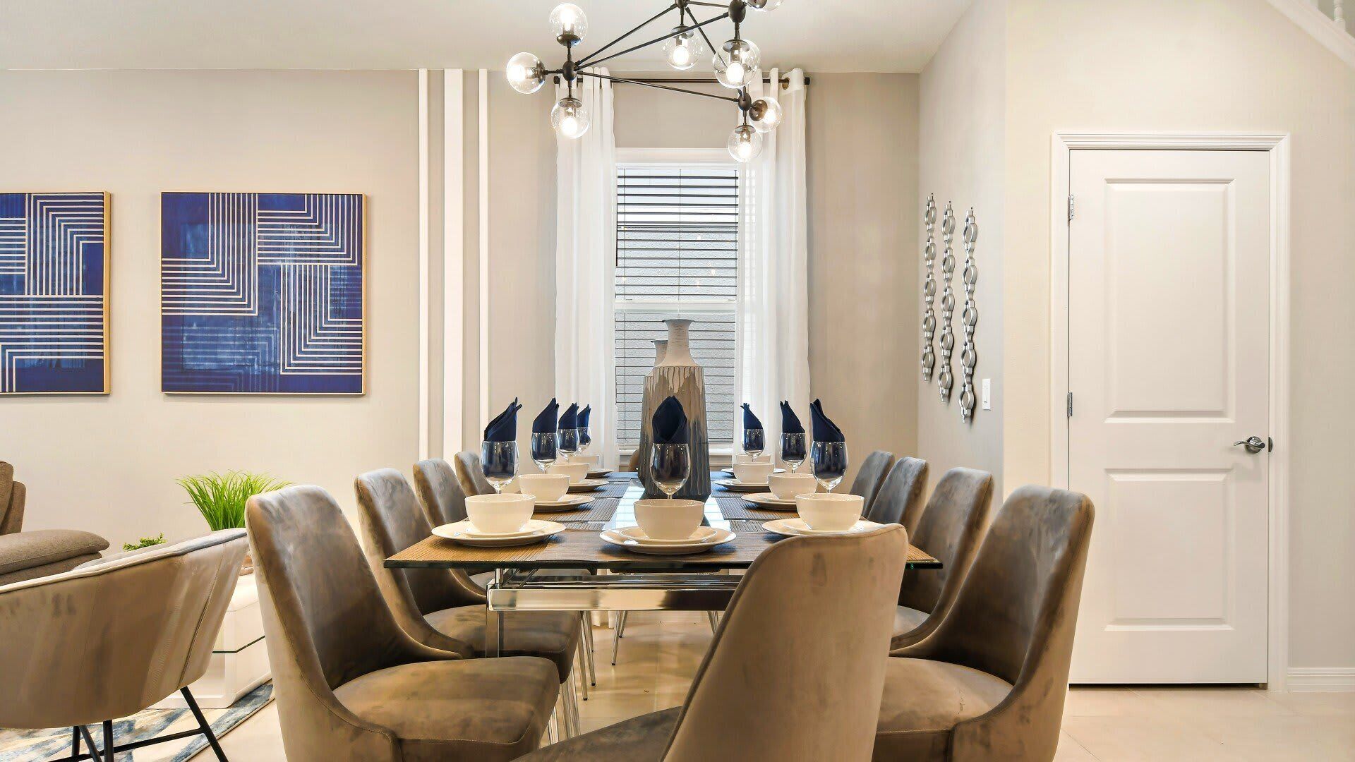 Ein Esszimmer mit einem großen Tisch, gedeckt für sechs Personen, umgeben von sechs gepolsterten Stühlen. An der Wand befinden sich zwei abstrakte Gemälde, auf dem Tisch eine große Vase und eine Tür mit dekorativer Griff.