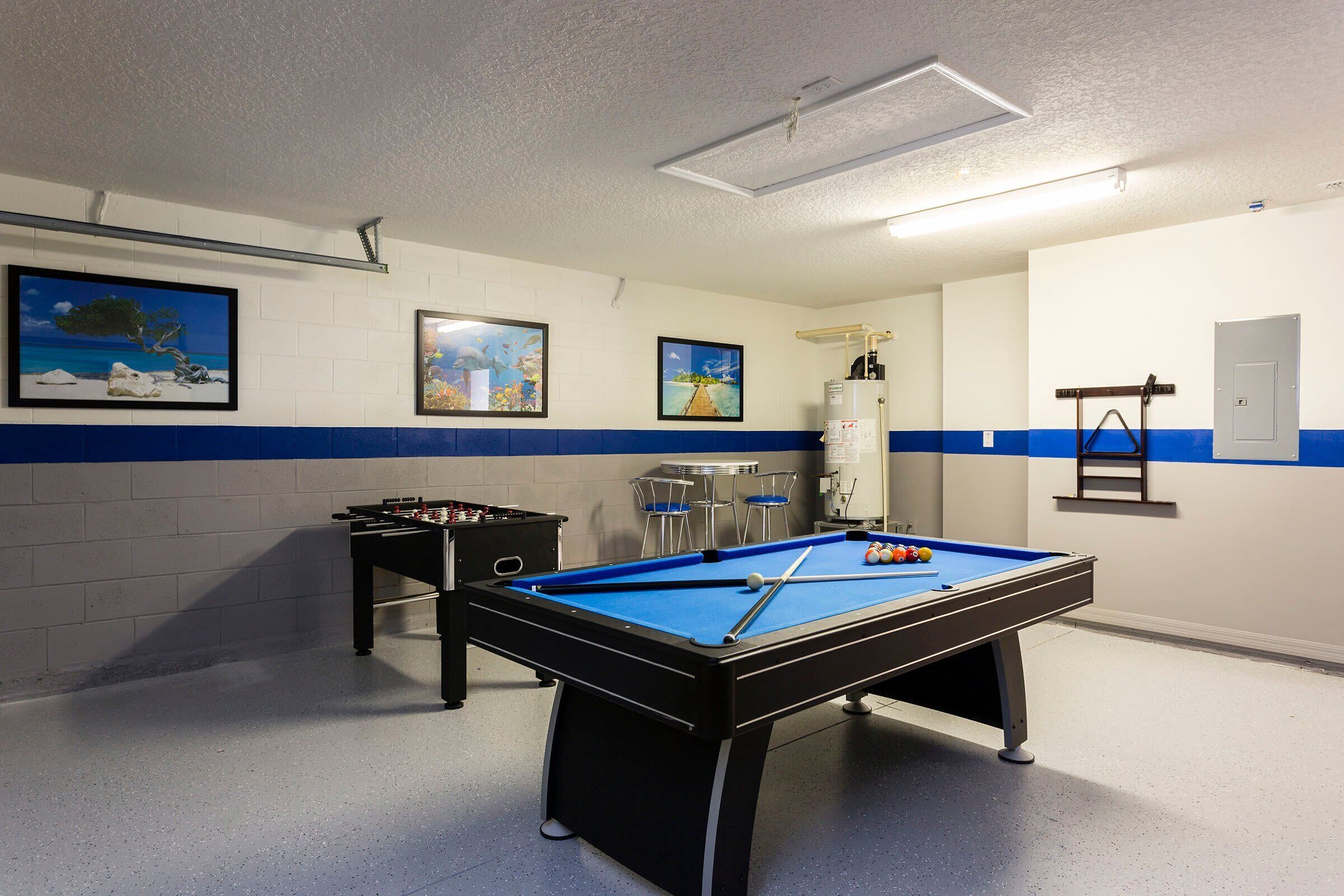 Een pooltafel met blauwe viltkleed, een tafelvoetbalspel en drie ingelijste foto's aan de muur.