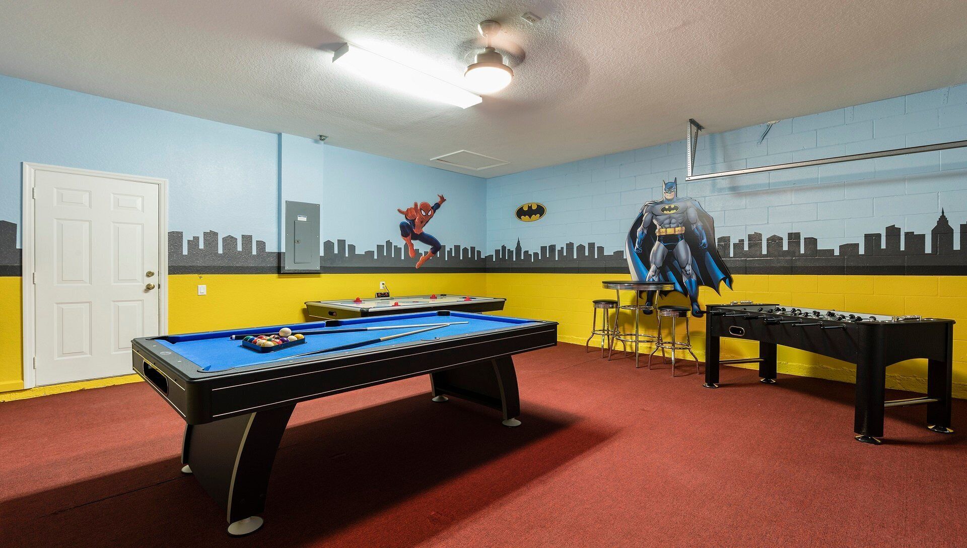 A room with a pool table, a foosball table, and a mural of superheroes on the wall.