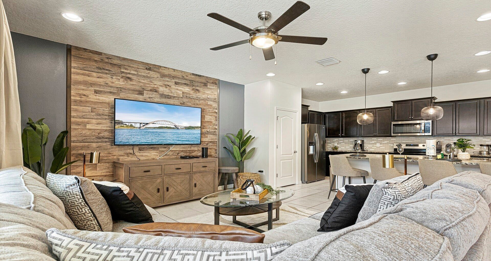 A modern living room with a large TV, comfortable seating, a ceiling fan, and a kitchen area with dark cabinets and stainless steel appliances.