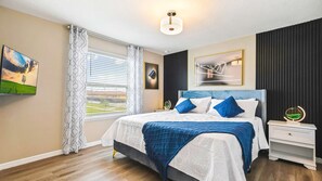 A bedroom with a bed, a bedside table, a nightstand, a window with curtains, and a wall-mounted light fixture. - Windsor At Westside 8BR Pool Spa Near Disney 2301 (Kissimmee)