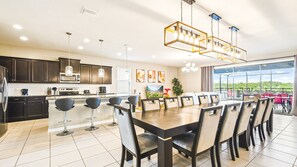 A modern kitchen with dark cabinets, a large island, and a dining area with a large table and chairs. - Windsor At Westside 8BR Pool Spa Near Disney 2301 (Kissimmee)