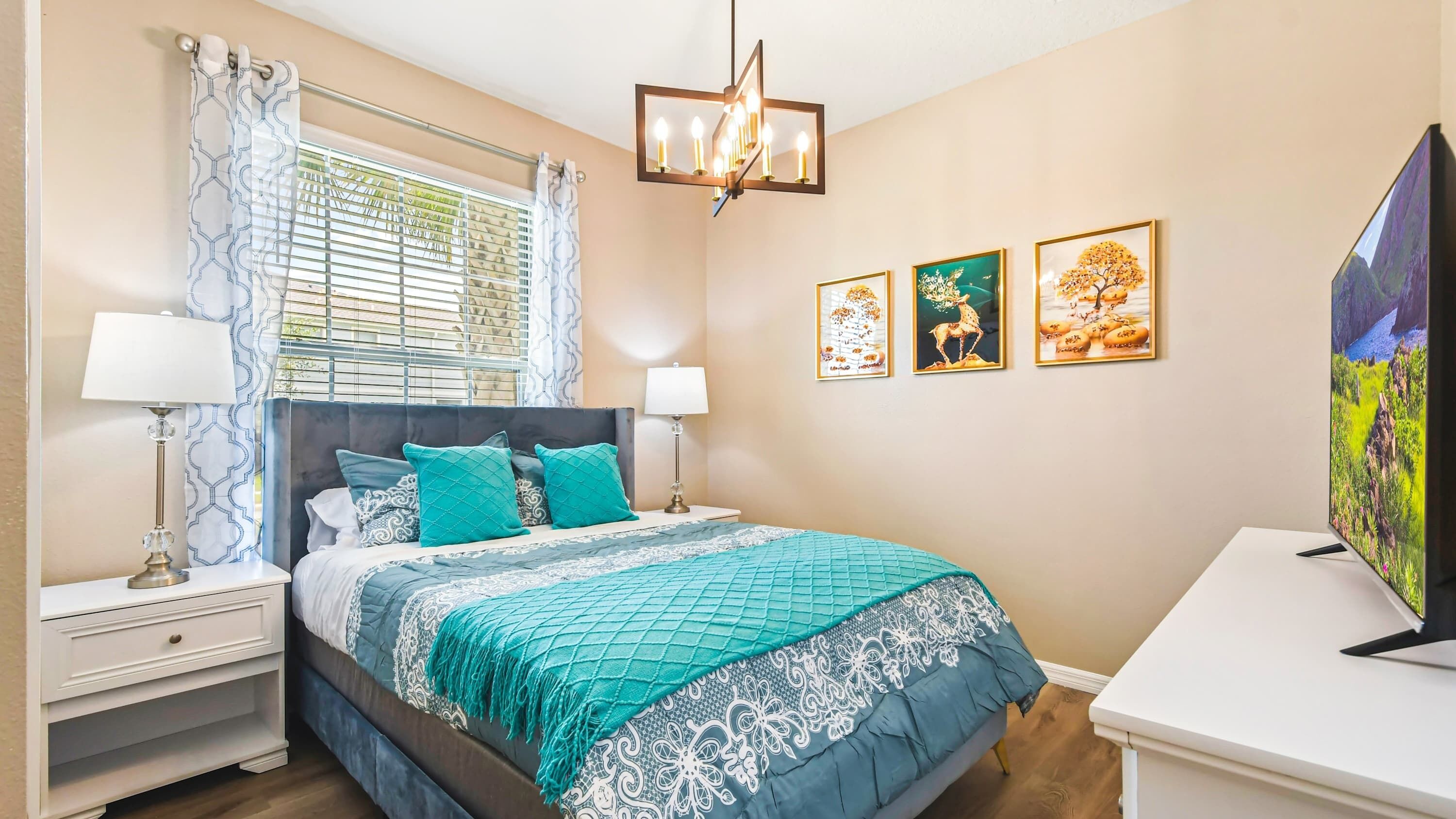 A bedroom with a bed, two bedside tables, a window with blinds, a chandelier, and three framed pictures on the wall.