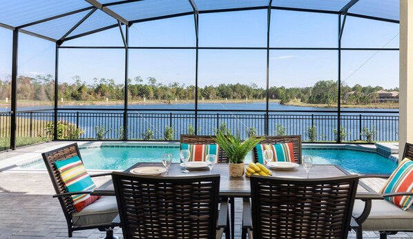 A covered outdoor area with a pool, dining table, and chairs, overlooking a lake. - Windsor At Westside 8BR Pool Spa Near Disney 2301 (Kissimmee)