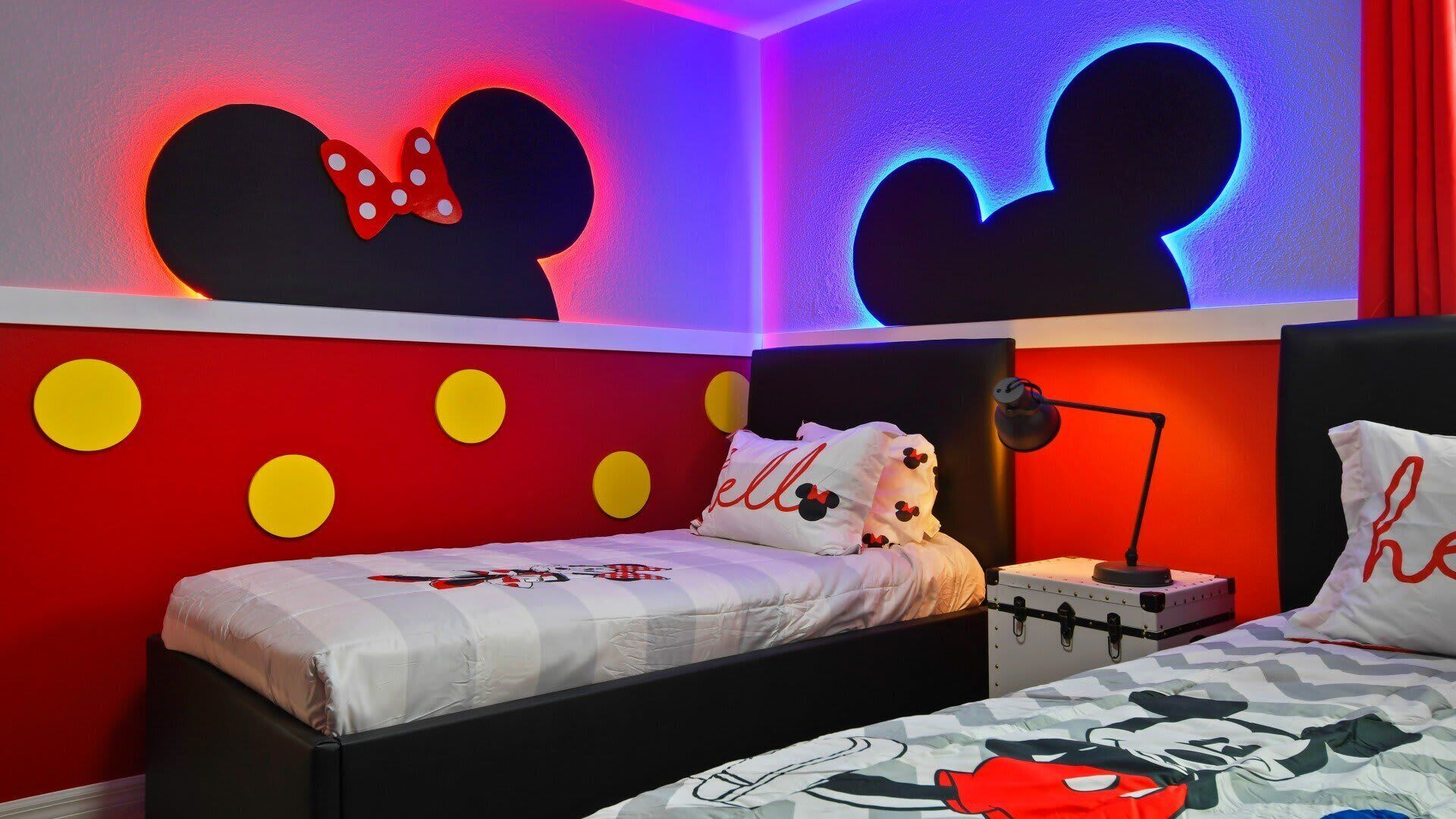 Et værelse med to senge, hvoraf den ene har et dynedække og puder med Mickey Mouse tema, og den anden et andet design. Væggene er dekoreret med store Mickey Mouse silhuetter og gule prikker.
