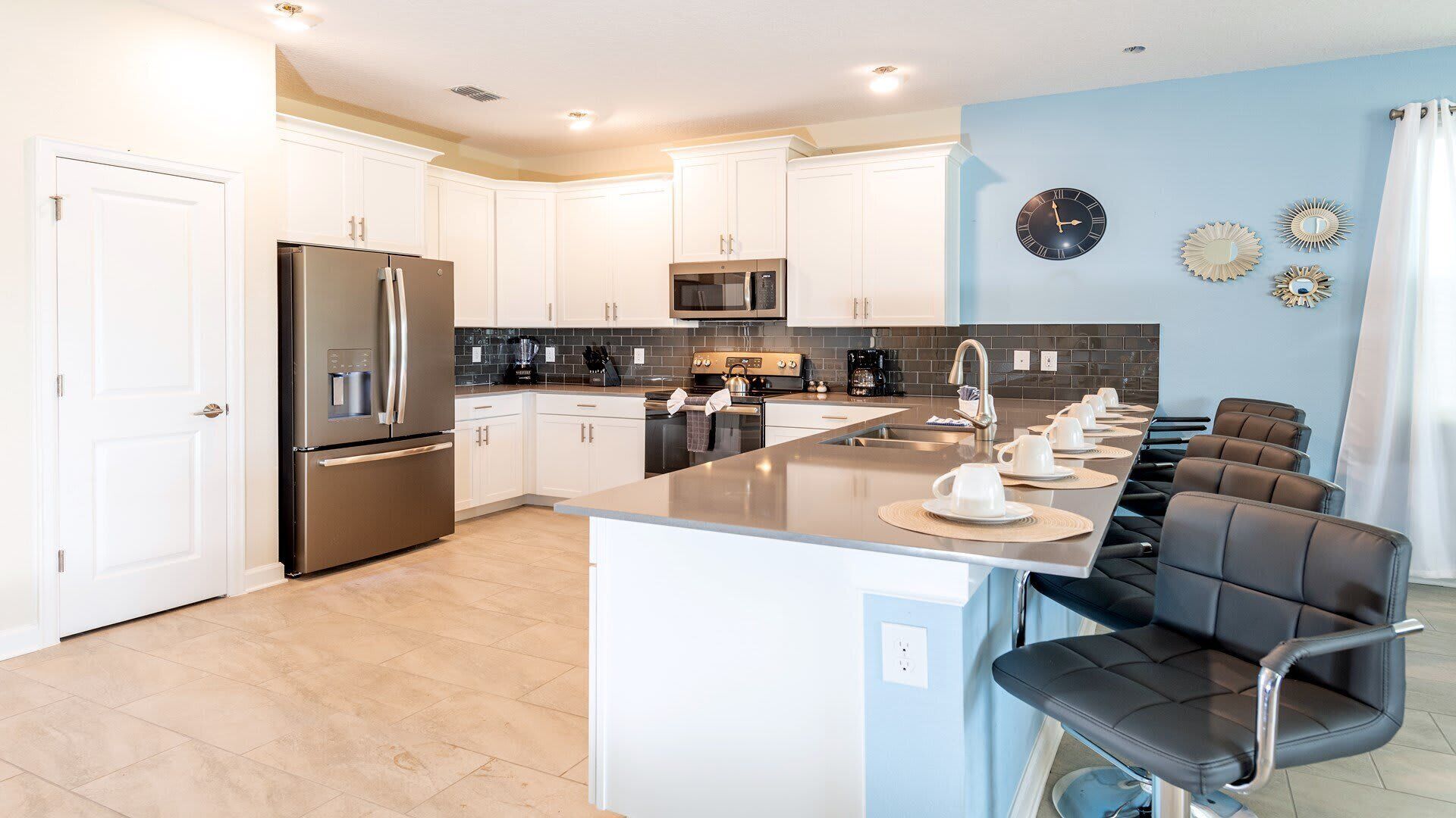 A modern kitchen with white cabinets, stainless steel appliances, a central island with seating, and decorative wall clocks.