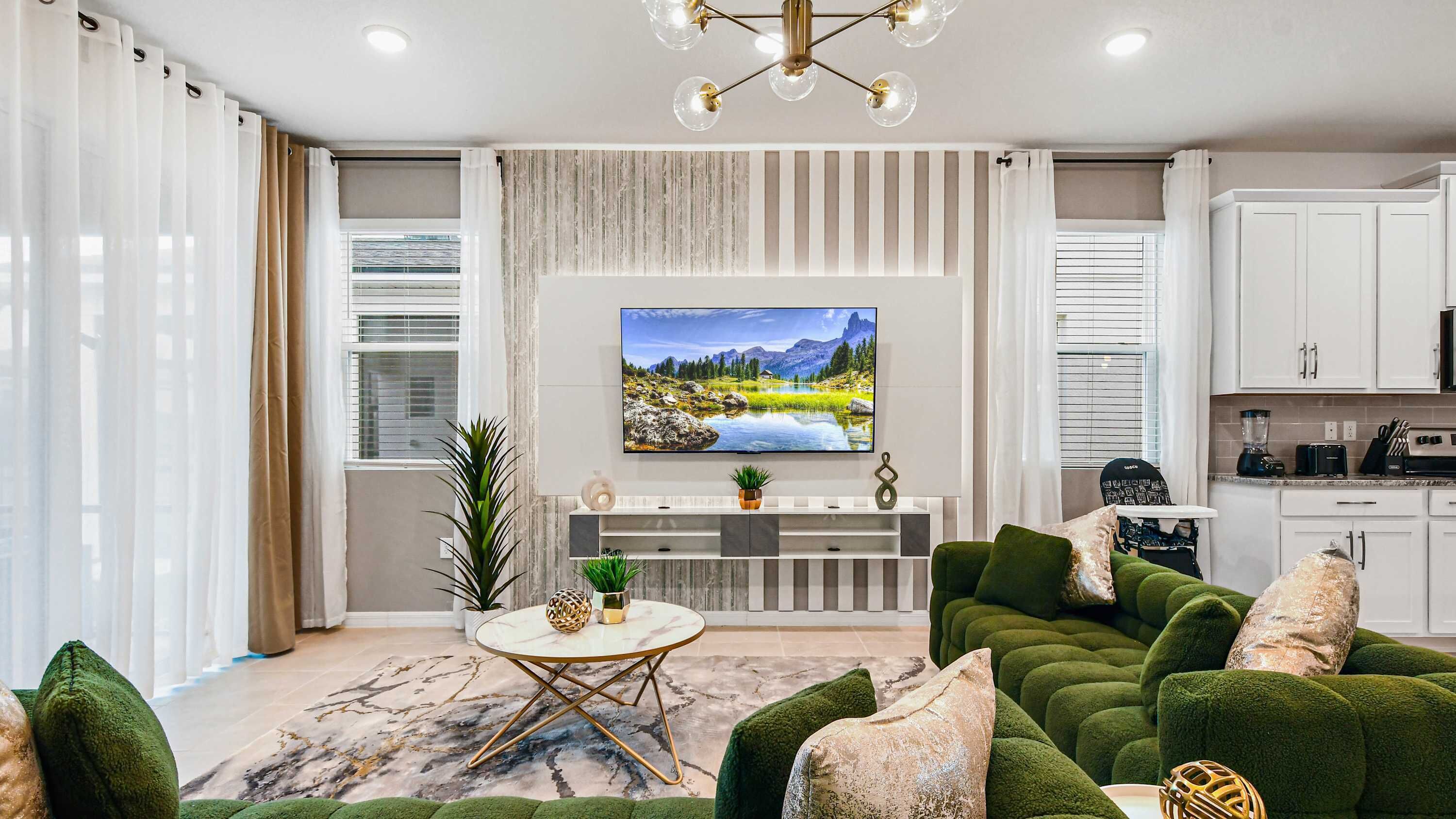 A modern living room with a large flat-screen TV, green upholstered sofas, a marble-patterned rug, and a kitchen area with white cabinets and appliances.