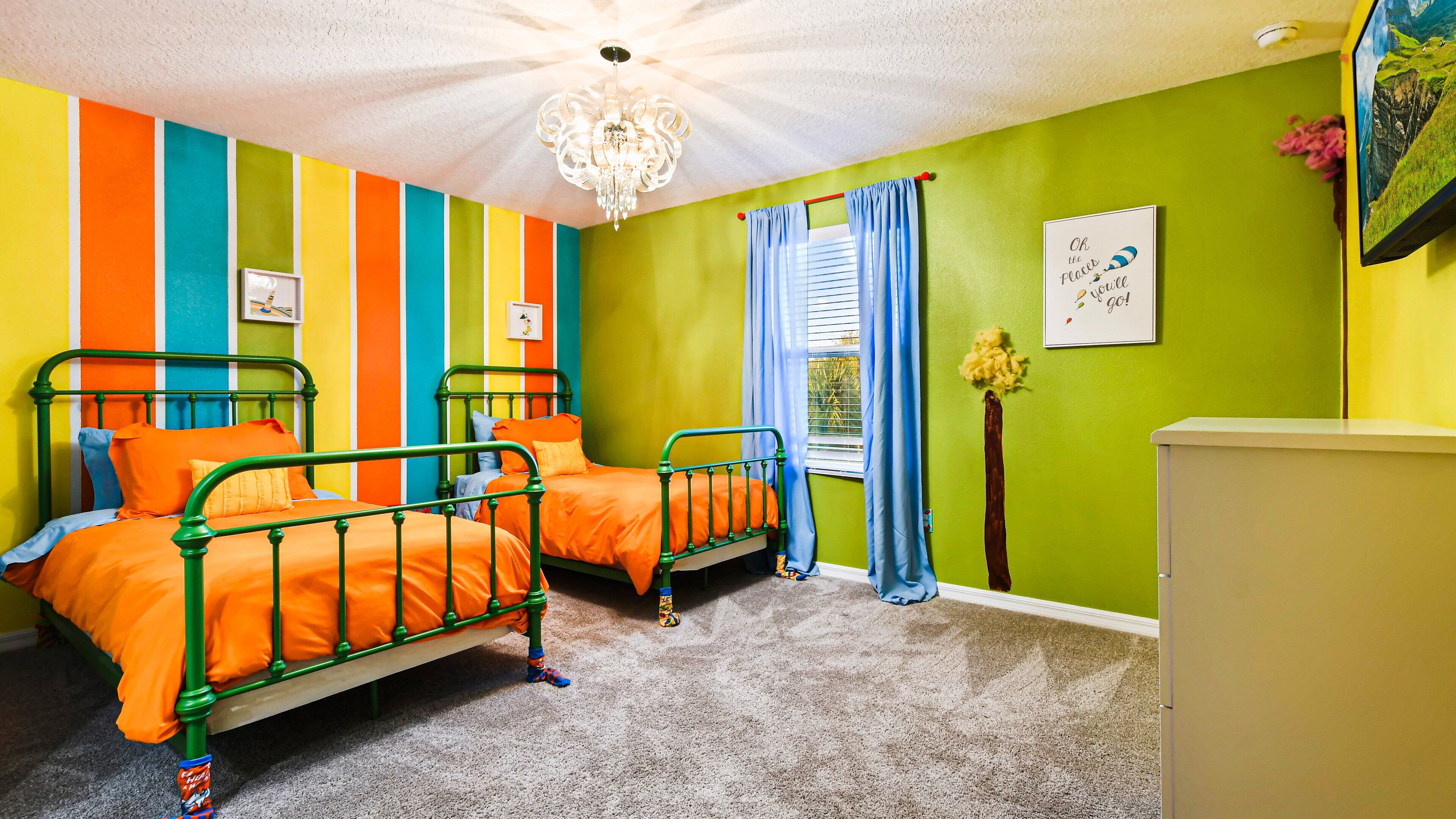 Quarto com duas camas de metal verde, paredes coloridas listradas, um candelabro e uma parede verde com uma moldura.