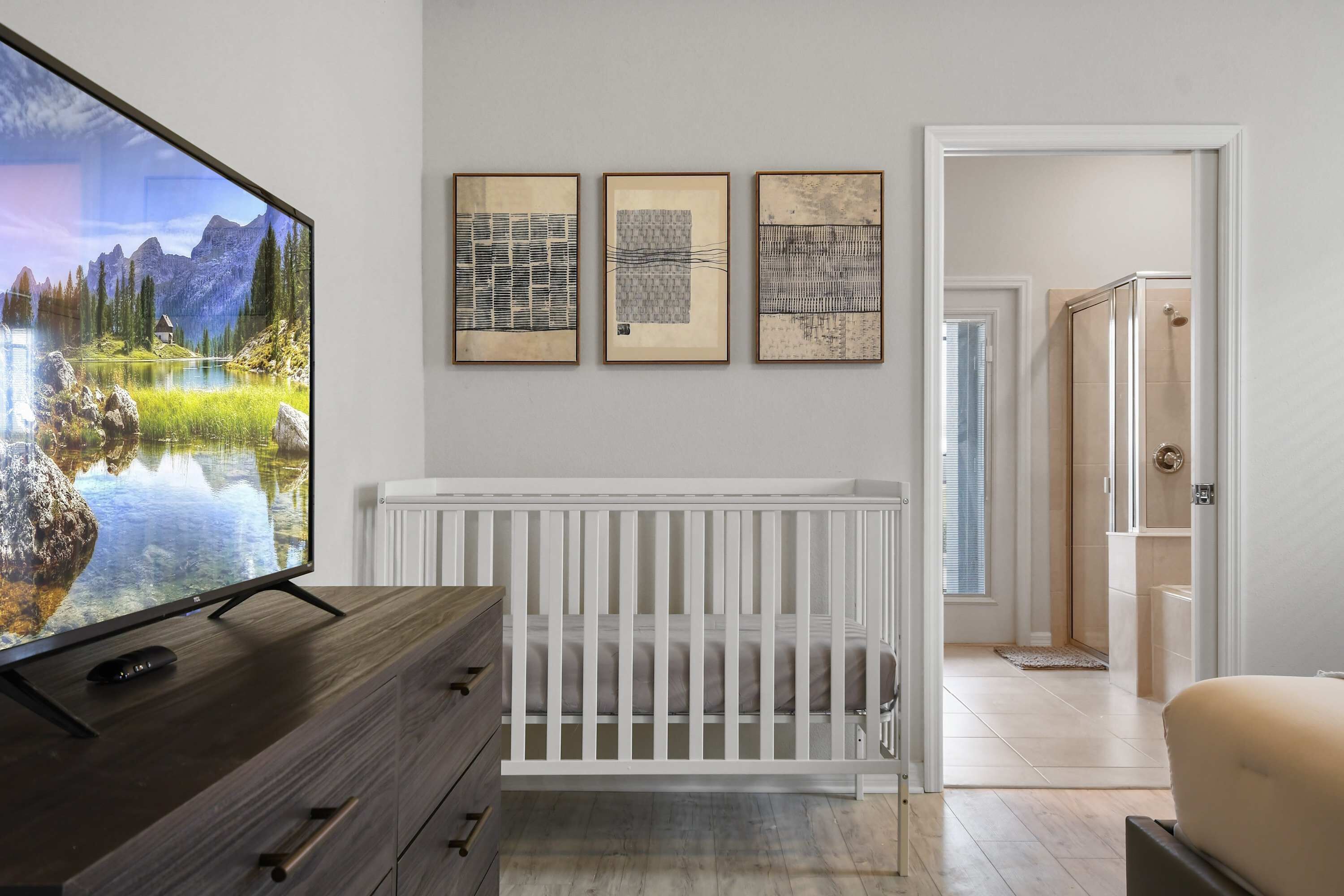 A modern room with a white crib, a flat-screen TV on a wooden dresser, and three framed pictures on the wall.