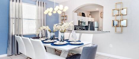 A dining area with a white table, blue placemats, white chairs, and a vase of white flowers.