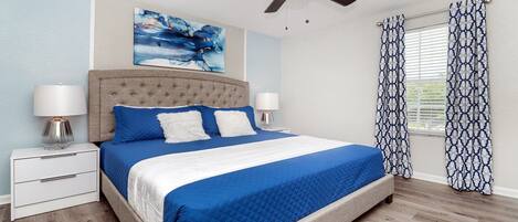 A bedroom with a large bed, two bedside tables, a ceiling fan, and a window with patterned curtains.