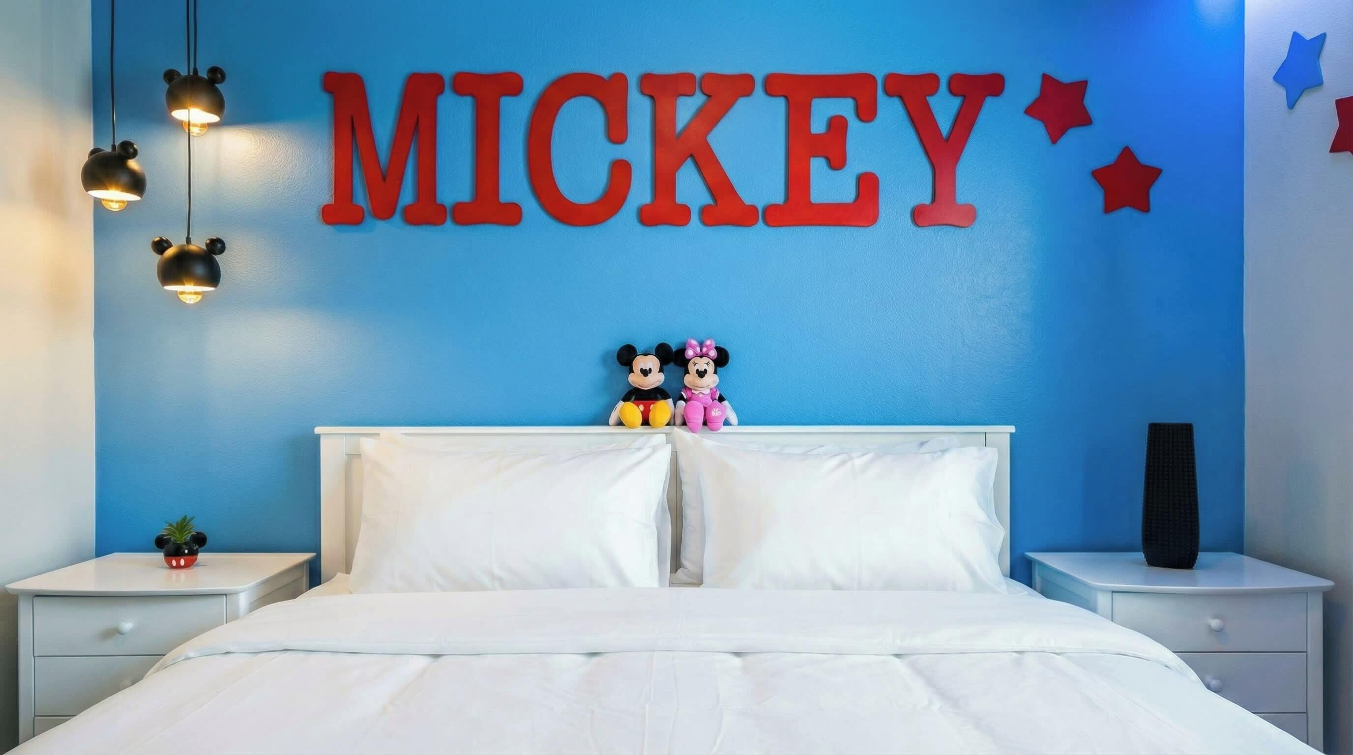 Een slaapkamer met een blauwe wand, een bed met wit beddengoed, pluchen Mickey en Minnie Mouse speelgoedfiguren op het hoofdeinde, en een groot bord met de tekst 'MICKEY' erboven.