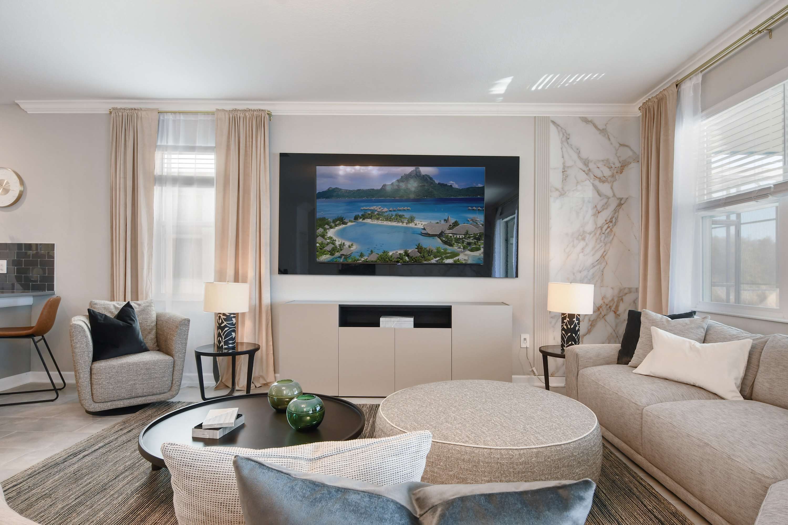 A modern hotel room with a large flat-screen TV mounted on the wall, a sofa, a round coffee table, and a dining area with a chair and a small table.
