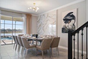 Uma sala de jantar com uma mesa grande posta para seis pessoas, rodeada por cadeiras estofadas, uma parede de mármore e um quadro de um pássaro.